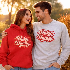 May include: A red hoodie and a gray sweatshirt with the text "Buby Bunny" in a retro font. The hoodie has a front pocket and a drawstring hood. The sweatshirt has a crew neck. The couple is smiling.