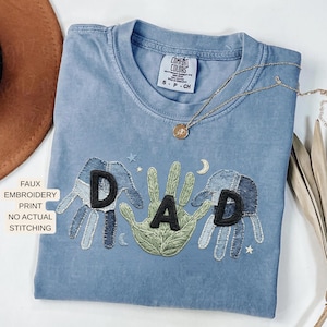 May include: A light blue Comfort Colors t-shirt with a faux embroidery print. The design features the word "DAD" with hand-shaped elements and a crescent moon. A gold necklace with a pendant is draped across the shirt.