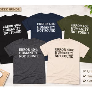May include: Five t-shirts in various colors, including black, navy, and olive green, each with the text "ERROR 404: HUMANITY NOT FOUND" in white. The shirts are displayed on a flat surface with a laptop and keyboard visible.