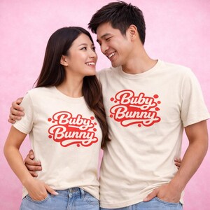 May include: Two people wearing matching cream-colored t-shirts with the red text "Buby Bunny". The text is in a retro, cursive font with small red dots. The background is a soft pink.
