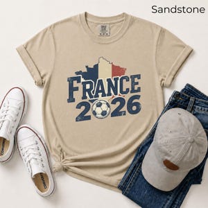France 2026 soccer shirt with a vintage map of France in French flag colors. Sandstone Comfort Colors t-shirt mockup with retro distressed typography and a soccer ball graphic. Trendy world cup football fan gear flat lay with sneakers.