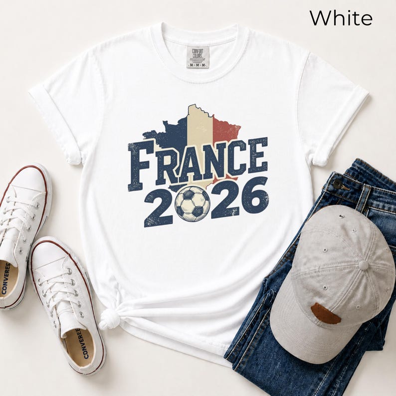 France 2026 soccer shirt with a vintage map of France in French flag colors. White Comfort Colors t-shirt mockup with retro distressed typography and a soccer ball graphic. Trendy world cup football fan gear flat lay with sneakers.