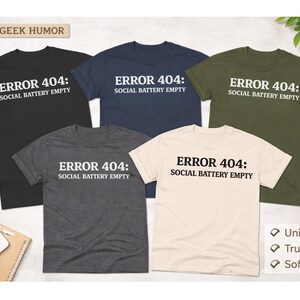 May include: Five t-shirts in black, navy, olive green, charcoal gray, and cream, each with the text "ERROR 404: SOCIAL BATTERY EMPTY" in white. The shirts are displayed on a white surface with a laptop, keyboard, and coffee cup.