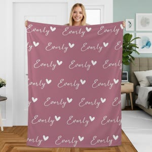 Puede incluir: Una manta rosa polvorienta con un patrón repetido del nombre "Everly" en escritura cursiva, acompañado de símbolos de corazón blanco. La manta se muestra, destacando su diseño y color.
