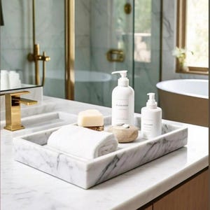 Può includere: Un set da toeletta per il bagno in marmo con un vassoio rettangolare che contiene un asciugamano bianco arrotolato, sapone e due flaconi con pompa bianchi. Il rubinetto e gli accessori della doccia sono dorati.