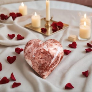Könnte beinhalten: Ein herzförmiges Deko-Objekt mit einem marmorierten rot-weißen Muster liegt auf einer weißen Oberfläche, umgeben von roten herzförmigen Blütenblättern und brennenden Kerzen. Ein goldener Tablett hält Kerzen, und eine Vase mit einer roten Rose ist sichtbar.
