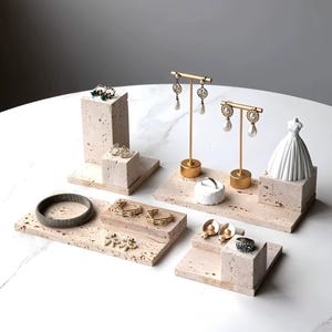 May include: A display of jewelry on textured, light-colored stone platforms. Gold-toned earring stands, a white dress form, and various rings, earrings, and a bracelet are arranged on the platforms. The jewelry includes pearls and gemstones.