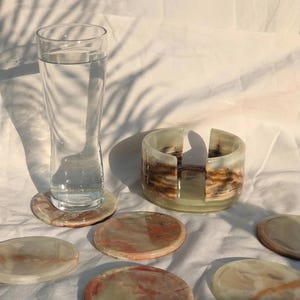Op de afbeelding: Een hoog glas water en verschillende ronde onderzetters van doorschijnend, gemarmerd steen. Er is ook een bijpassende onderzetterhouder aanwezig. De onderzetters hebben tinten roze, bruin en crème.