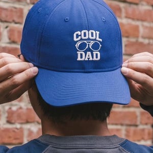 May include: A royal blue baseball cap with the words "COOL DAD" embroidered in white. The cap has a curved brim and a six-panel design. The text includes a pair of glasses. The cap is being held by a person.