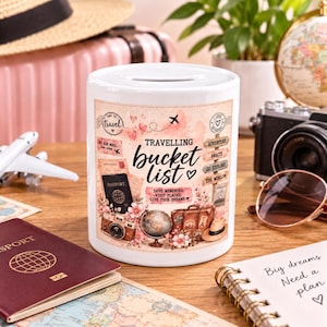 May include: A white ceramic money box featuring a travel-themed graphic with the text "TRAVELLING BUCKET LIST," "SAVE MEMORIES, VISIT PLACES, LIVE YOUR DREAMS," and "ADVENTURE AWAITS, GO EXPLORE, THE WORLD." The design includes a passport, camera, globe, and suitcase.