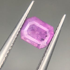 Könnte beinhalten: Ein facettierter, rechteckig geschliffener, rosa Edelstein, der von einer silbernen Pinzette gehalten wird. Der Stein hat einen helleren rosa Innenraum und einen dunkleren rosa Außenrand. Kleine dunkle Einschlüsse sind sichtbar. Die Pinzette ist diagonal vor grauem Hintergrund.