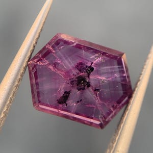 Puede incluir: Una gema facetada, hexagonal, de un profundo color magenta. La gema tiene vetas internas e inclusiones oscuras, sostenida por unas pinzas plateadas. Las facetas de la piedra capturan la luz, mostrando su claridad y color.