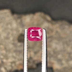 May include: A square-cut ruby gemstone, held by silver tweezers. The ruby is a rich, deep red colour and is faceted to enhance its brilliance. The background is a blurred, neutral tone.