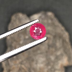 Könnte beinhalten: Ein facettierter, runder, leuchtend roter Edelstein, der von einer silbernen Pinzette gehalten wird. Der Edelstein hat ein sternförmiges Muster in der Mitte. Der Hintergrund ist verschwommen und neutral gefärbt.