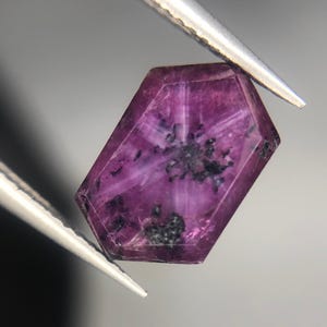 Op de afbeelding: Een gefacetteerde, zeshoekige edelsteen in magenta. De steen, vastgehouden door een pincet, vertoont een stervormig patroon met donkere insluitsels. De facetten benadrukken de natuurlijke schoonheid van dit mineraal.