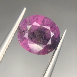 Puede incluir: Una gema rosa facetada, de forma ovalada, sujeta con pinzas. La gema tiene una estructura interna compleja con inclusiones oscuras y una superficie brillante y multifacética. El color es un rosa intenso y saturado.