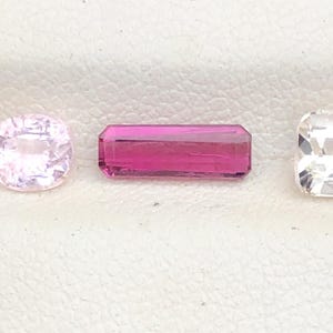 May include: Three gemstones of different shapes and colors are displayed. A light pink, round gemstone is on the left. A rectangular, magenta gemstone is in the center. A square, white gemstone is on the right.