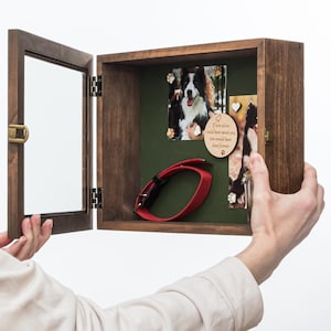 Könnte beinhalten: Eine offene, dunkelbraune Holz-Gedenkbox für Haustiere mit Glastür, die ein rotes Hundehalsband, Fotos und ein herzförmiges Holzornament zeigt. Die Box hat einen Messingverschluss.