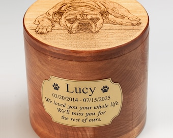 May include: A wooden pet urn with a light brown lid and a darker brown body. The lid features an engraved image of a dog and the name "Lucy". A gold-colored plaque on the urn reads "Lucy" and includes dates and a memorial message.