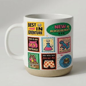 May include: White ceramic mug with a speckled finish and a tan base. The mug is decorated with colorful sticker-like designs, including text that reads "Best in Adventure" and "New Achievement!"