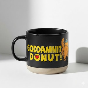 Puede incluir: Taza de cerámica negra con el texto "GODDAMNIT DONUT!" en amarillo, un corazón rojo y una ilustración de dibujos animados de un perro con corona. La parte inferior de la taza es beige.