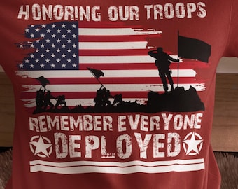 May include: Maroon t-shirt with the text "HONORING OUR TROOPS" above a graphic of the American flag and silhouettes of soldiers. Below the flag, the text reads "REMEMBER EVERYONE DEPLOYED" with star accents. The shirt is a crew neck.