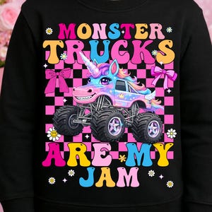 May include: Black sweatshirt featuring a colorful graphic of a monster truck with a unicorn horn, set against a pink and black checkered background. The text "MONSTER TRUCKS ARE MY JAM" is displayed in vibrant colors.