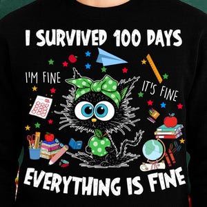 以下が含まれることがあります： 「I Survived 100 Days」と「Everything is Fine」の文字が入った黒いスウェットシャツ。緑のリボンと水玉模様の漫画の猫が中央にあり、学校をテーマにしたグラフィックが周りに描かれています。