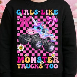 May include: Black sweatshirt with a colorful graphic of a unicorn monster truck. The text reads "GIRLS LIKE MONSTER TRUCKS TOO" in a playful font, with pink bows and daisy accents. The background is a pink and black checkered pattern.