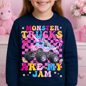 May include: Navy blue sweatshirt with a colorful graphic of a monster truck designed as a unicorn. The text reads "MONSTER TRUCKS ARE MY JAM" in a playful font, with floral and bow accents. The design is set against a pink and black checkered background.