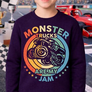 May include: A dark purple sweatshirt featuring a colorful graphic of a monster truck inside a circle with the words "MONSTER TRUCKS ARE MY JAM". The design uses orange, yellow, green, and blue colors. Stars surround the bottom of the graphic.