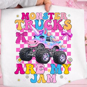 May include: A white graphic tee featuring a colorful monster truck designed as a unicorn. The text "MONSTER TRUCKS ARE MY JAM" is displayed in a retro font with pink, orange, yellow, and blue colors. The background is a pink and white checkered pattern.