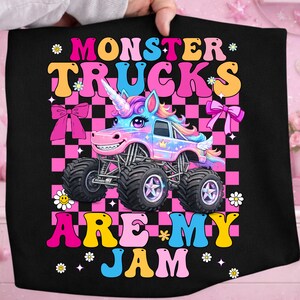 May include: Black graphic tee featuring a cartoon monster truck with a unicorn horn, set against a pink and black checkered background. The text reads "MONSTER TRUCKS ARE MY JAM" in colorful, playful lettering.