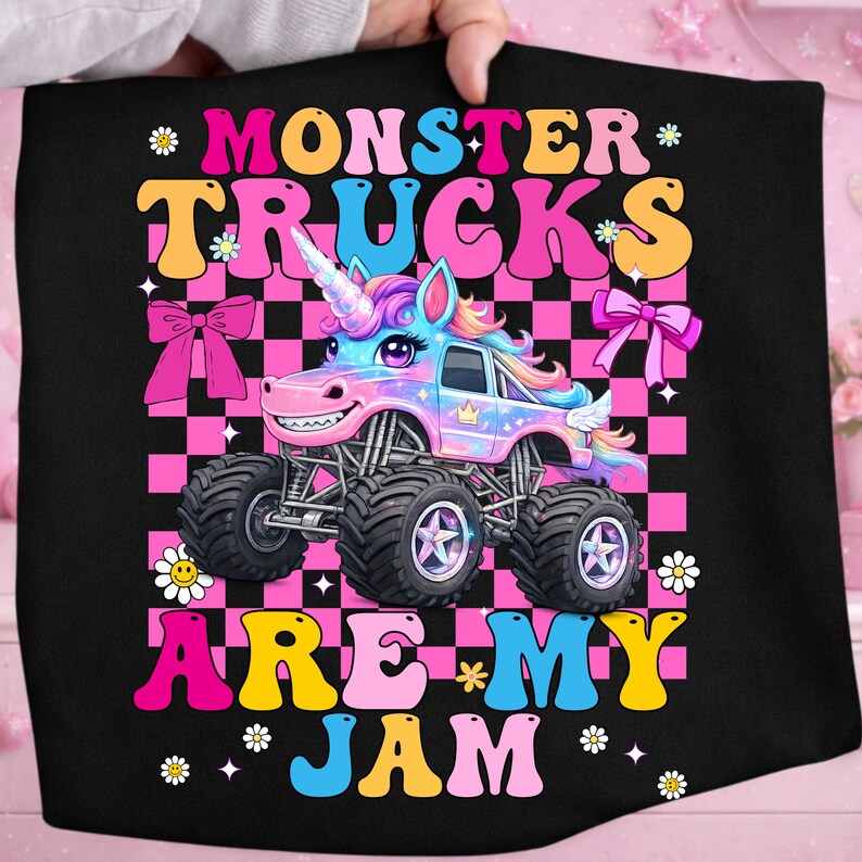 May include: Black graphic tee featuring a cartoon monster truck with a unicorn horn, set against a pink and black checkered background. The text reads "MONSTER TRUCKS ARE MY JAM" in colorful, playful lettering.