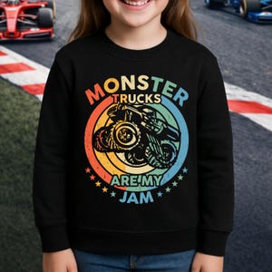 May include: Black sweatshirt featuring a colorful graphic of a monster truck inside a circular design with the text "MONSTER TRUCKS ARE MY JAM". The design includes orange, yellow, blue, and green colors. The sweatshirt is worn by a person.