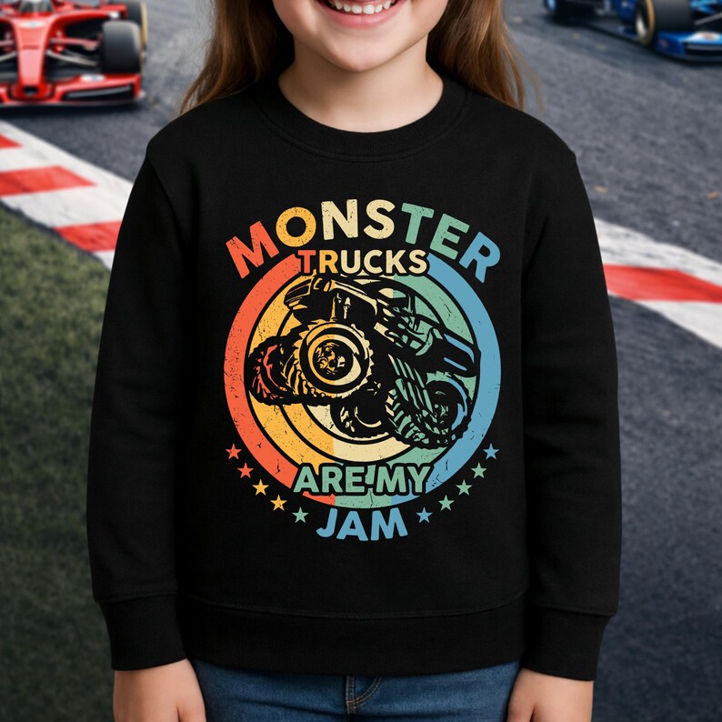 May include: Black sweatshirt featuring a colorful graphic of a monster truck inside a circular design with the text "MONSTER TRUCKS ARE MY JAM". The design includes orange, yellow, blue, and green colors. The sweatshirt is worn by a person.