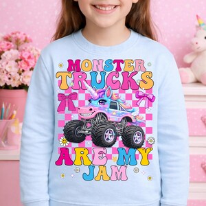 May include: Light blue sweatshirt with a colorful graphic of a unicorn monster truck. The text "MONSTER TRUCKS ARE MY JAM" is displayed above and below the truck. The design includes pink bows, flowers, and a checkered background.