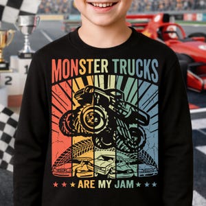 May include: Black sweatshirt with a colorful graphic that says "MONSTER TRUCKS ARE MY JAM." The design features a monster truck jumping, with tire tracks and car silhouettes below. The background has a retro sunset design.