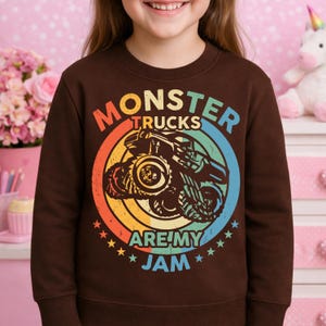 May include: A brown sweatshirt featuring a colorful graphic of a monster truck inside a circular design. The text "MONSTER TRUCKS ARE MY JAM" is printed around the truck. The design includes orange, yellow, blue, and green colors.