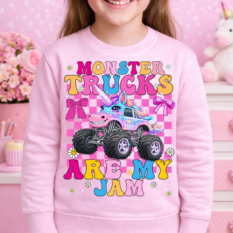 May include: Pink sweatshirt featuring a colorful monster truck designed as a unicorn. The text "MONSTER TRUCKS ARE MY JAM" is displayed in a playful font. The design includes bows, flowers, and a checkered background.