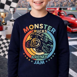 May include: Navy blue sweatshirt with a colorful graphic of a monster truck inside a circle. The text "MONSTER TRUCKS ARE MY JAM" is displayed in a rainbow of colors. The design includes stars and a vintage style.