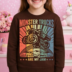 May include: A brown sweatshirt featuring a colorful graphic of a monster truck with the text "MONSTER TRUCKS ARE MY JAM." The design includes a monster truck, tire tracks, and several cars in a retro style.