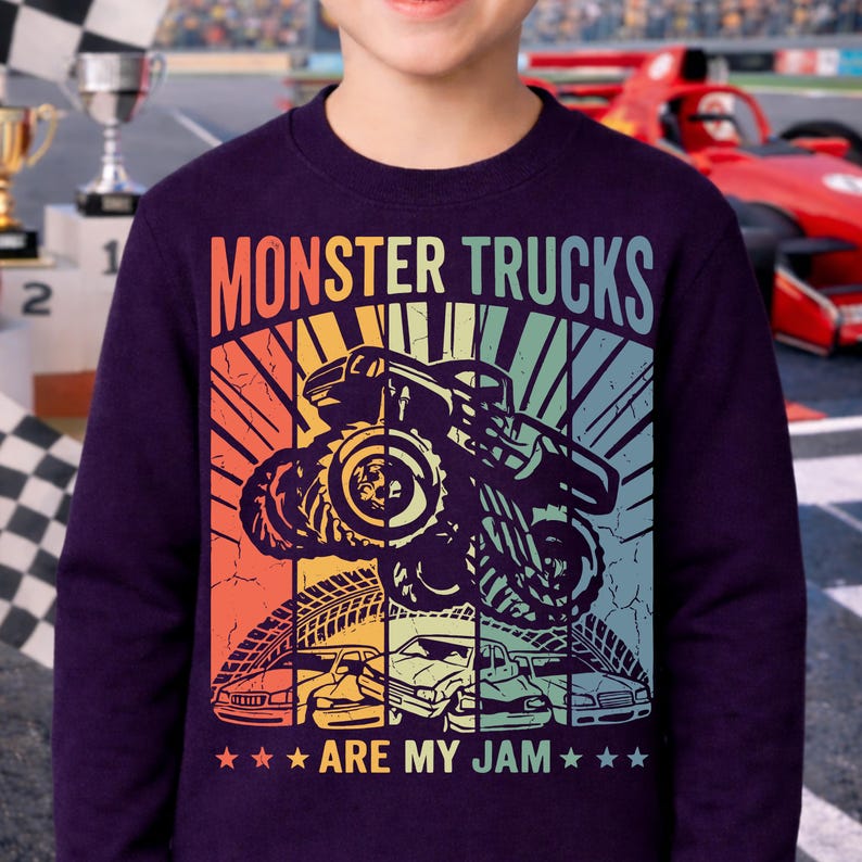 May include: A dark purple sweatshirt featuring a graphic of a monster truck jumping over cars. The text "MONSTER TRUCKS ARE MY JAM" is displayed in a retro style with a colorful gradient background. The design includes tire tracks and stars.