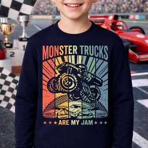 May include: Navy blue sweatshirt with a colorful graphic featuring a monster truck, the text "MONSTER TRUCKS" and "ARE MY JAM". The design includes a retro sunset and several cars.