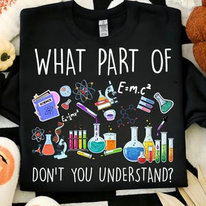 Może przedstawiać: Czarna bluza z okrągłym dekoltem z białym napisem "WHAT PART OF DON'T YOU UNDERSTAND?". Projekt zawiera ilustracje naukowe, takie jak zlewki, prob&oacute;wki i modele atomowe.