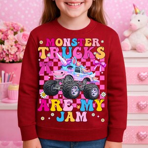 May include: A maroon sweatshirt featuring a colorful graphic of a monster truck with a unicorn horn. The text reads "MONSTER TRUCKS ARE MY JAM" in yellow, pink, and blue. The design includes bows and flowers.
