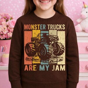 Puede incluir: Una sudadera marr&oacute;n con un gr&aacute;fico de monster truck y el texto "MONSTER TRUCKS ARE MY JAM". El dise&ntilde;o incluye una ilustraci&oacute;n de monster truck en negro, azul, amarillo, naranja y beige.