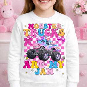 May include: White sweatshirt featuring a colorful graphic of a monster truck designed as a unicorn. The text "MONSTER TRUCKS ARE MY JAM" is displayed in vibrant colors. The design includes bows, flowers, and a checkered background.