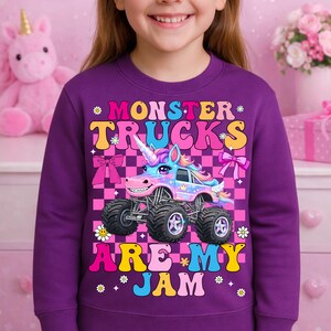 May include: A purple sweatshirt featuring a colorful graphic of a monster truck designed as a unicorn. The text "MONSTER TRUCKS ARE MY JAM" is displayed above and below the truck. The background is a pink and black checkered pattern.