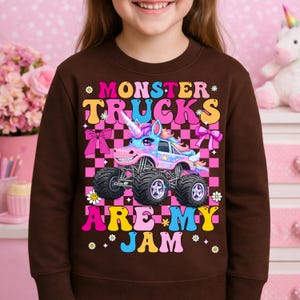 May include: A brown sweatshirt featuring a colorful graphic of a monster truck designed as a unicorn. The text "MONSTER TRUCKS ARE MY JAM" is displayed in a playful font. The background is a pink and black checkered pattern.
